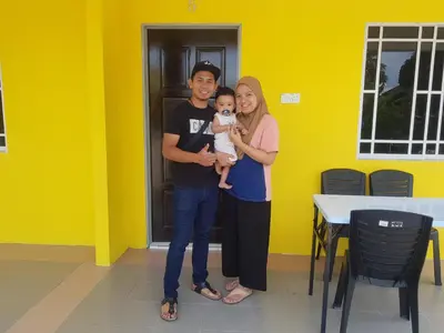 a man and woman holding a baby in front of a yellow wall at Room/Homestay NazaDja in Sungai Besar
